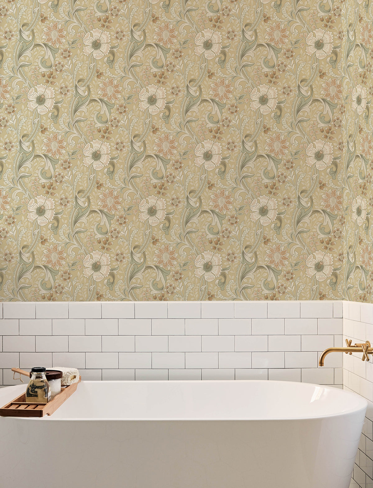 A white bathtub with a gold faucet is set against a wall adorned with York Wallcoverings Chesapeake Liora Floral Trail Wallpaper and white subway tiles. A wooden bath tray holds a candle, jar, and soap.
