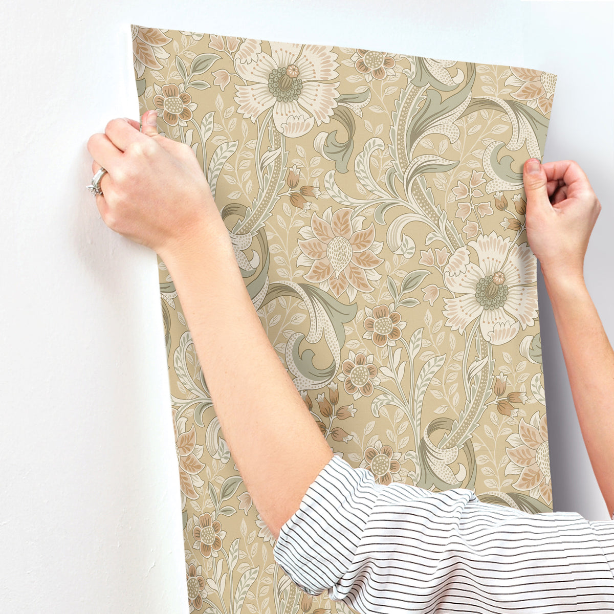 A person wearing a striped shirt applies York Wallcoverings Chesapeake Liora Floral Trail Wallpaper, featuring beige, green, and cream tones, to a white wall.