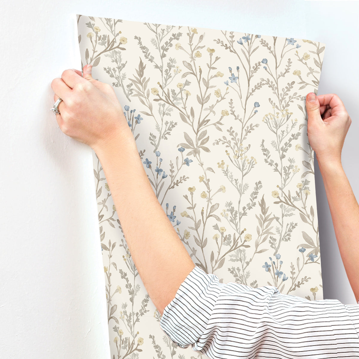 Wearing a striped shirt and a ring, a person applies York Wallcoverings’ Chesapeake Laverne Wildflowers Wallpaper—featuring a delicate floral pattern in soft beige, blue, and yellow tones—to a white wall; only their hands and arms are visible.
