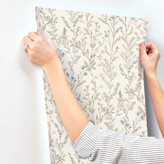 A person wearing a striped shirt and a ring on their left hand is applying Chesapeake Laverne Wildflowers Wallpaper by York Wallcoverings, featuring beige, blue, and yellow small flowers and leaves, to a white wall. Only their arms and hands are visible.