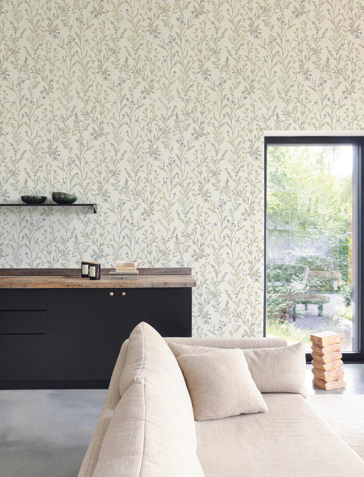 A cozy living room features a beige sofa, black cabinets, and a wooden countertop. Wall shelves hold bowls above the Chesapeake Laverne Wildflowers Wallpaper by York Wallcoverings, while a large window overlooks a green garden. A Jenga game sits on the floor near the window.