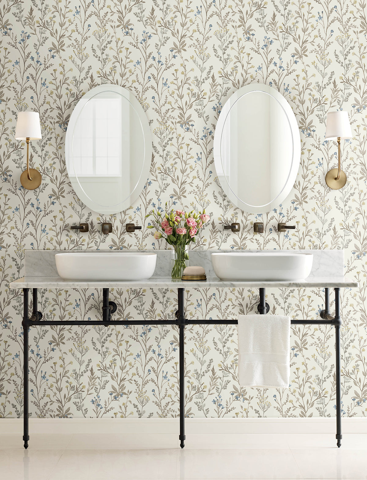 A bathroom featuring two round mirrors, white sinks, wall-mounted faucets, Chesapeake Laverne Wildflowers Wallpaper by York Wallcoverings, two wall sconces, a vase of flowers, and a towel hanging on a rack below the counter.