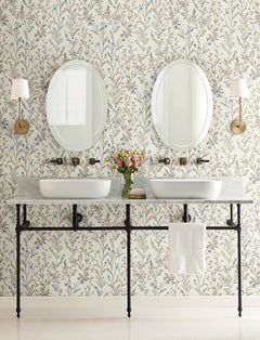 A bathroom featuring two round mirrors, white sinks, wall-mounted faucets, Chesapeake Laverne Wildflowers Wallpaper by York Wallcoverings, two wall sconces, a vase of flowers, and a towel hanging on a rack below the counter.