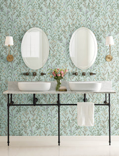 Double sink bathroom vanity with a marble countertop, two round mirrors, wall-mounted faucets, brass sconces, a white towel, and a vase of flowers set against York Wallcoverings’ Chesapeake Laverne Wildflowers Wallpaper.