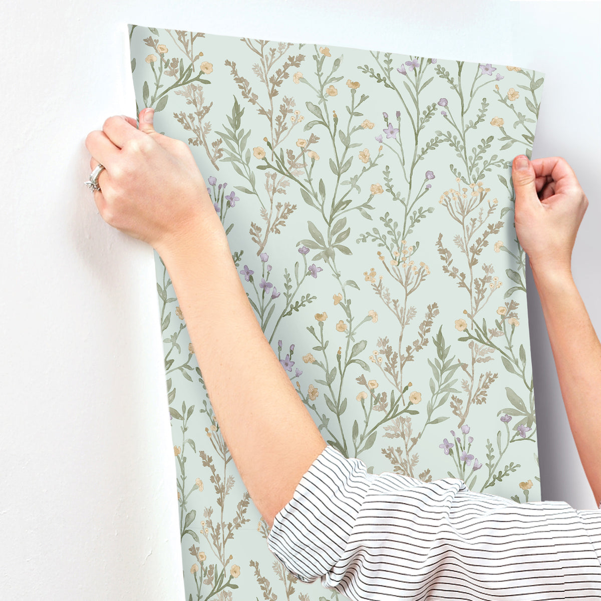 Wearing a striped shirt, a person hangs the Chesapeake Laverne Wildflowers Wallpaper by York Wallcoverings—featuring a light green background and delicate, leafy plant designs—on a white wall.