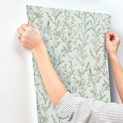 Wearing a striped shirt, a person hangs the Chesapeake Laverne Wildflowers Wallpaper by York Wallcoverings—featuring a light green background and delicate, leafy plant designs—on a white wall.