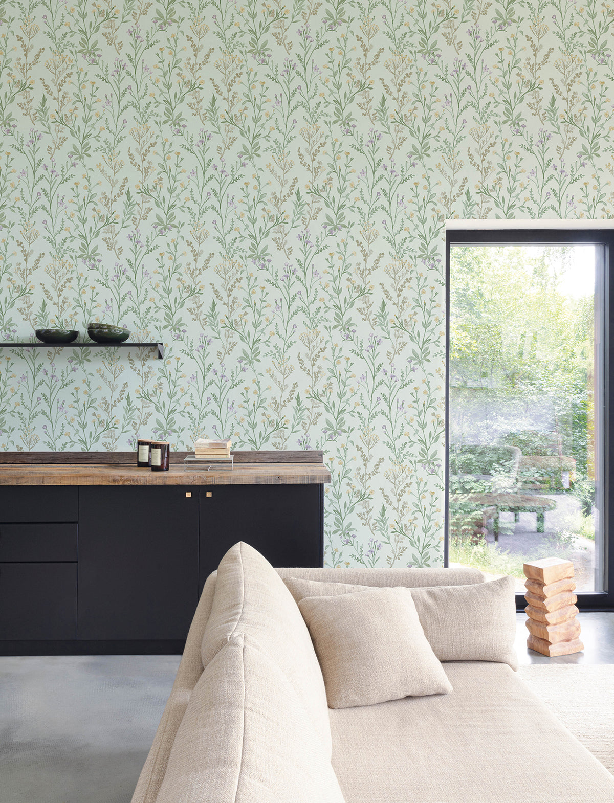 A modern living room featuring a beige sofa, black cabinets, wooden countertop, and Chesapeake Laverne Wildflowers Wallpaper by York Wallcoverings. A window reveals a lush garden outside, while Jenga blocks are stacked on the floor near the sofa.