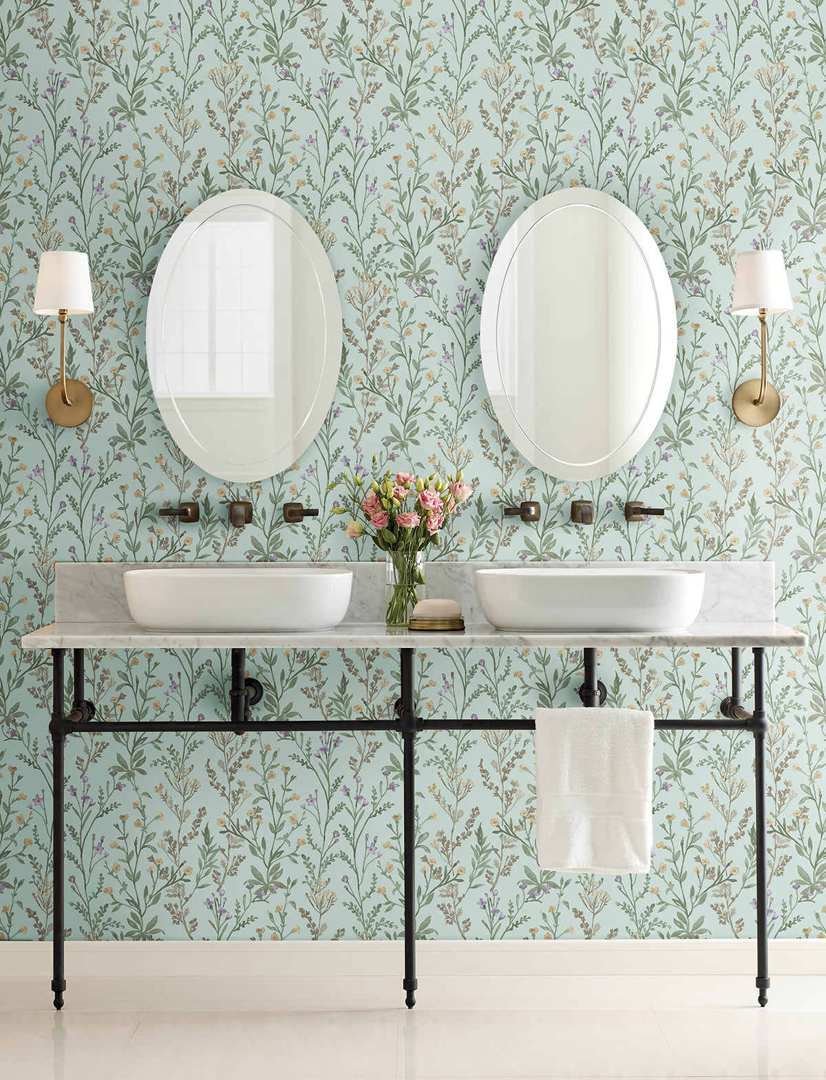 Double bathroom vanity with two round mirrors, white vessel sinks, a marble countertop, brass fixtures, Chesapeake Laverne Wildflowers Wallpaper by York Wallcoverings as the backdrop, two wall sconces, a vase of flowers, and a white towel hanging from the counter.