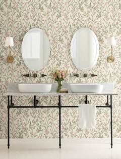 A bathroom featuring York Wallcoverings’ Chesapeake Laverne Wildflowers Wallpaper, two round mirrors above dual sinks, wall sconces, a shared marble countertop with a towel hanging on a metal rail, and a vase of flowers placed in the center.