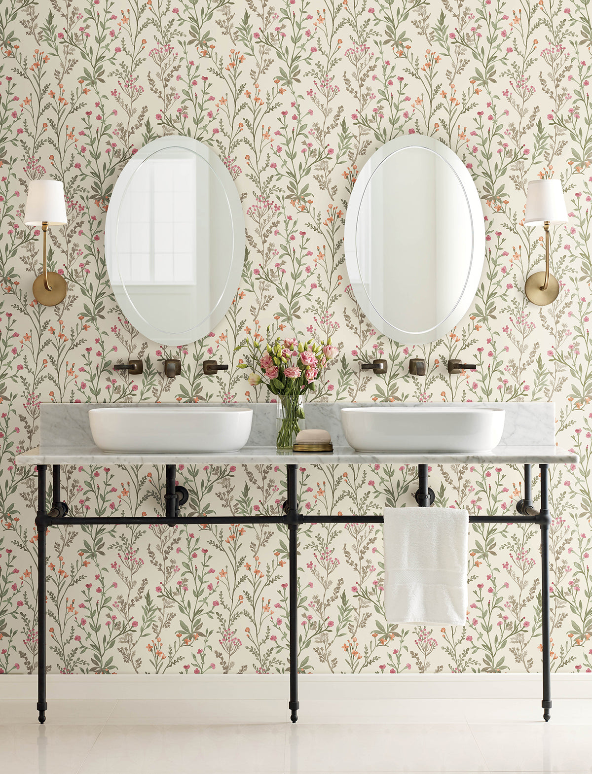 A bathroom featuring Chesapeake Laverne Wildflowers Wallpaper by York Wallcoverings, two round mirrors, wall sconces, twin sinks set in a marble counter, and a vase of flowers in the center. A towel hangs beneath the counter.