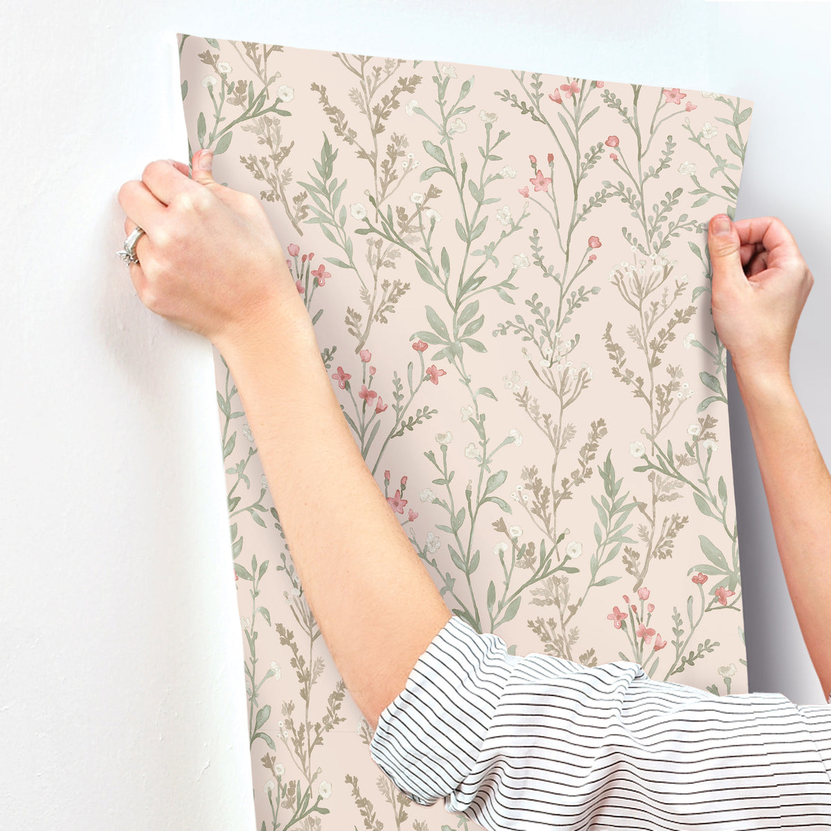 A person in a striped shirt aligns Chesapeake Laverne Wildflowers Wallpaper by York Wallcoverings, featuring pink and green botanical patterns, onto a white wall.