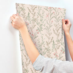 A person in a striped shirt aligns Chesapeake Laverne Wildflowers Wallpaper by York Wallcoverings, featuring pink and green botanical patterns, onto a white wall.