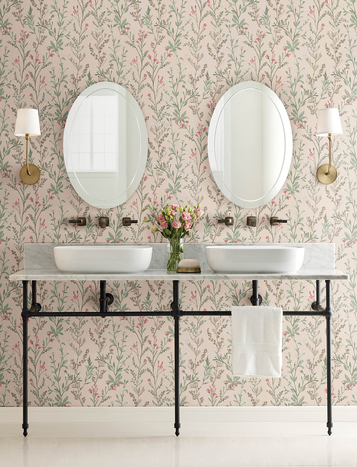 Two sinks with oval mirrors above, a marble countertop, a towel, wall-mounted lights, and a vase of flowers. The walls are adorned with Chesapeake Laverne Wildflowers Wallpaper by York Wallcoverings, featuring a charming pink and green floral pattern that enhances the classic and elegant style of the room.