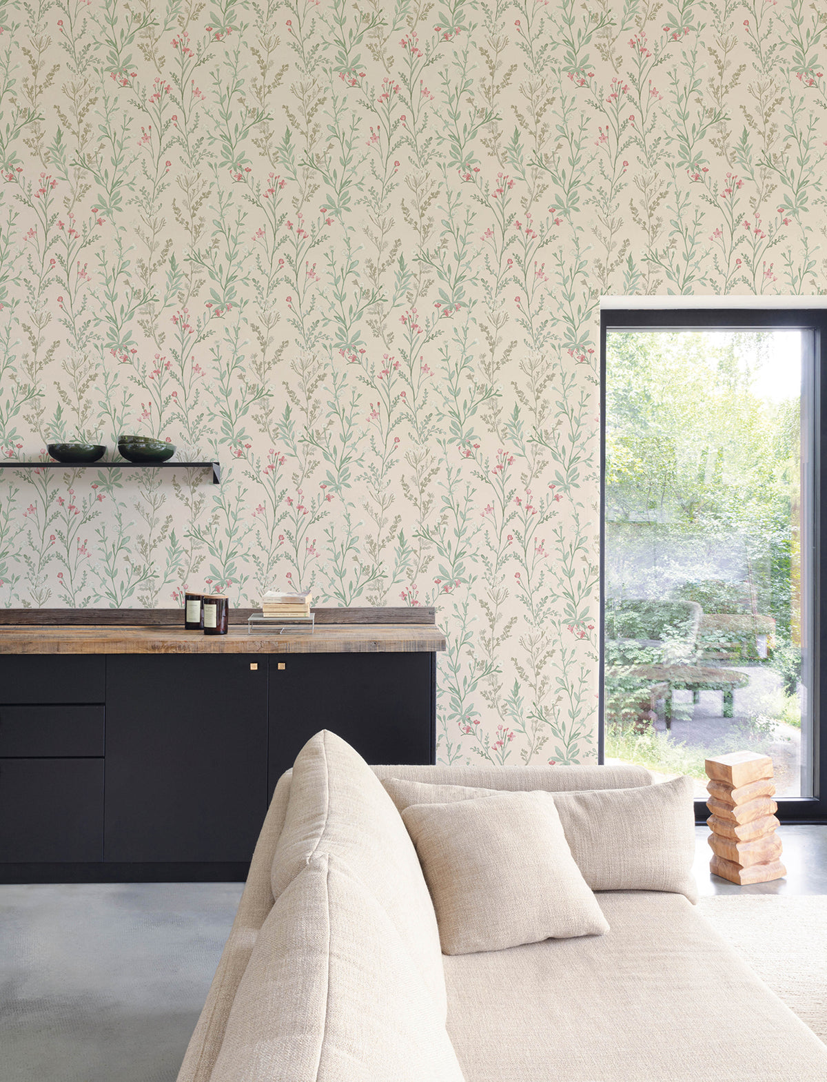 A modern living room featuring a beige sofa, black cabinets, a wooden countertop, and stacked books is brought to life with York Wallcoverings’ Chesapeake Laverne Wildflowers Wallpaper. A large window fills the cozy, stylish space with natural light and views of lush greenery outside.