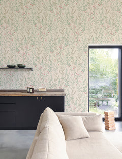 A modern living room featuring a beige sofa, black cabinets, a wooden countertop, and stacked books is brought to life with York Wallcoverings’ Chesapeake Laverne Wildflowers Wallpaper. A large window fills the cozy, stylish space with natural light and views of lush greenery outside.