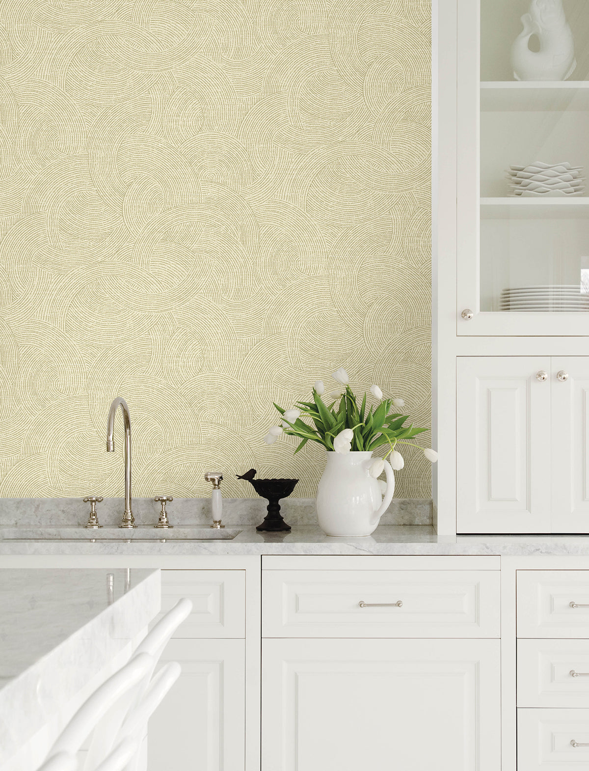 Modern white kitchen featuring marble countertops, a sink, and open shelves displaying plates. The walls are adorned with Chesapeake Harrison Swirling Geometric Wallpaper by York Wallcoverings in a light beige pattern. Beside the faucet sits a white pitcher with white tulips and a black decorative bird.