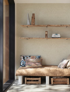 Cozy reading nook with a tan cushion, patterned pillows, and wooden crates beneath. Above, wooden shelves hold books and decor against a light wall adorned in Chesapeake Harrison Swirling Geometric Wallpaper by York Wallcoverings. Natural light streams in from a large window on the left.