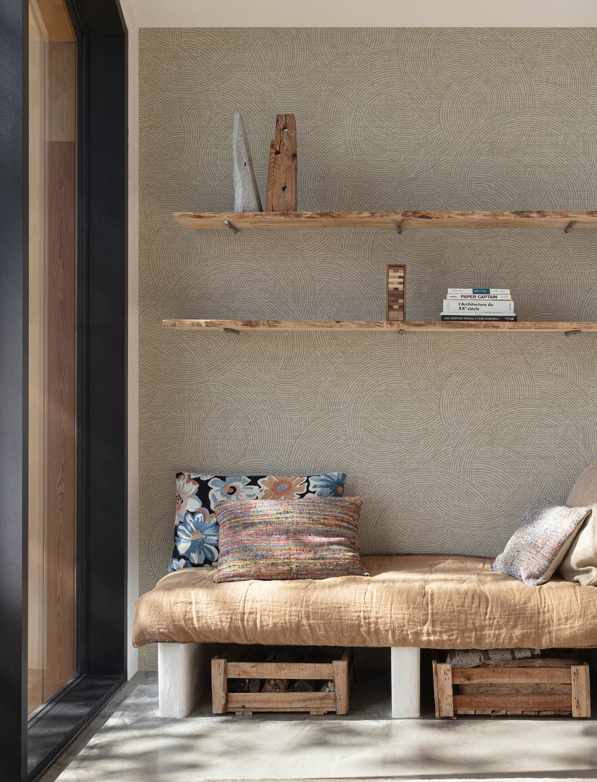 A cozy reading nook featuring a beige cushion, patterned pillows, and wooden shelves displaying books and decor is set against Chesapeake Harrison Swirling Geometric Wallpaper by York Wallcoverings. Wooden crates are neatly stored underneath the seat.