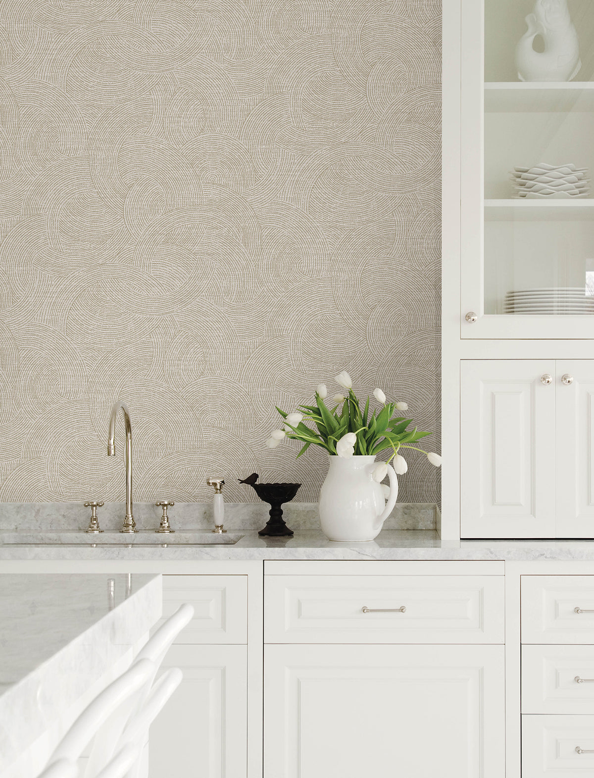 White kitchen with marble countertops, Chesapeake Harrison Swirling Geometric Wallpaper by York Wallcoverings, white cabinetry, a silver faucet, a white pitcher holding white tulips, and white dishes displayed on glass shelves.