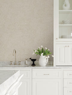 White kitchen with marble countertops, Chesapeake Harrison Swirling Geometric Wallpaper by York Wallcoverings, white cabinetry, a silver faucet, a white pitcher holding white tulips, and white dishes displayed on glass shelves.