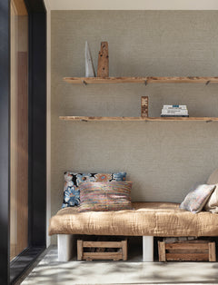 Create a cozy corner featuring Chesapeake Harrison Swirling Geometric Wallpaper by York Wallcoverings, a built-in bench topped with a tan blanket and colorful pillows. Wooden shelves above display books and decor, while storage crates sit below the bench. Sunlight pours in through a large window on the left, illuminating the inviting space.