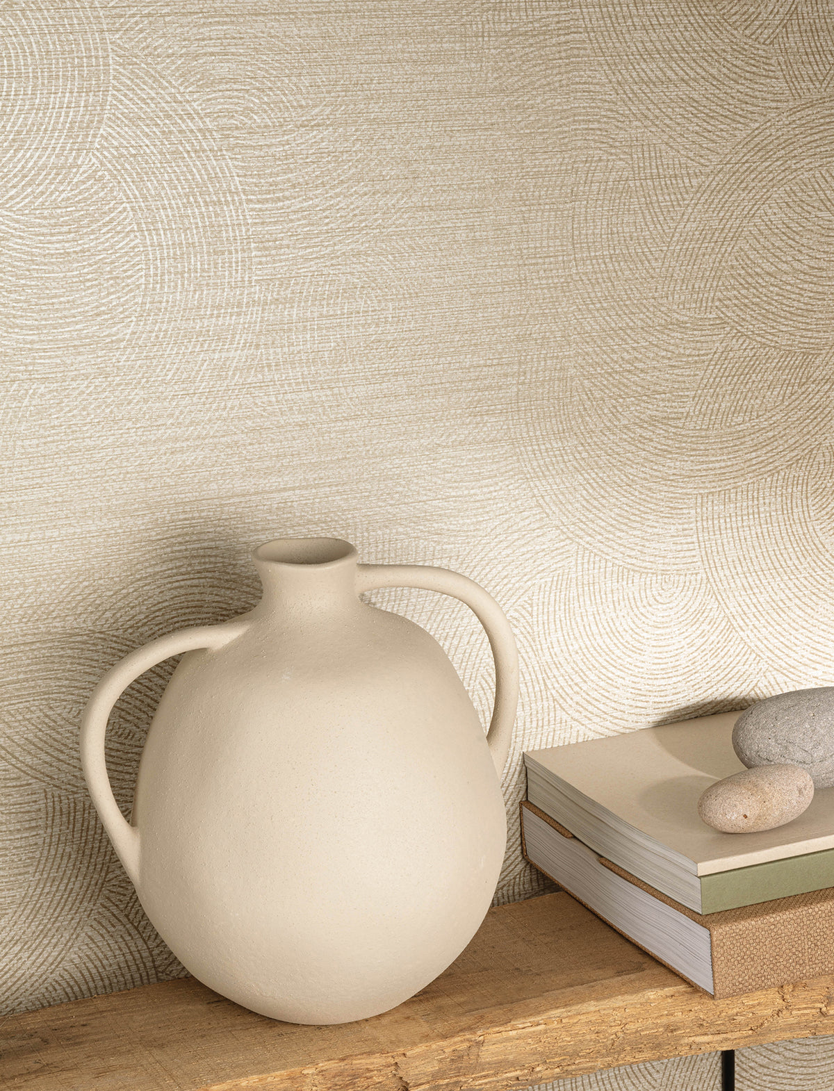 A round, cream-colored ceramic vase with two handles sits on a wooden shelf beside stacked books and two smooth stones, set against a wall adorned with Chesapeake Harrison Swirling Geometric Wallpaper by York Wallcoverings.
