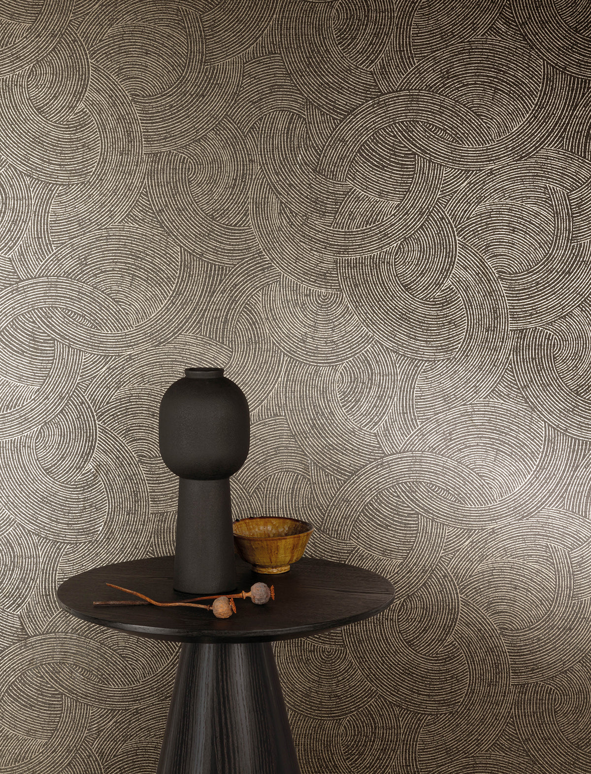 A round dark table with a tall matte black vase, a small wooden bowl, and dried plant stems is set in front of a wall adorned with Chesapeake Harrison Swirling Geometric Wallpaper by York Wallcoverings in beige and brown tones.