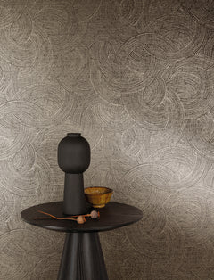 A round dark table with a tall matte black vase, a small wooden bowl, and dried plant stems is set in front of a wall adorned with Chesapeake Harrison Swirling Geometric Wallpaper by York Wallcoverings in beige and brown tones.