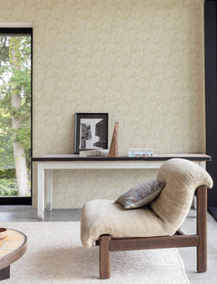A cozy beige lounge chair with a cushion sits near a slim console table displaying framed art, a box, and a wood sculpture, set against a wall adorned with York Wallcoverings’ Chesapeake Sonny Fronds Wallpaper beside a large window overlooking green trees.