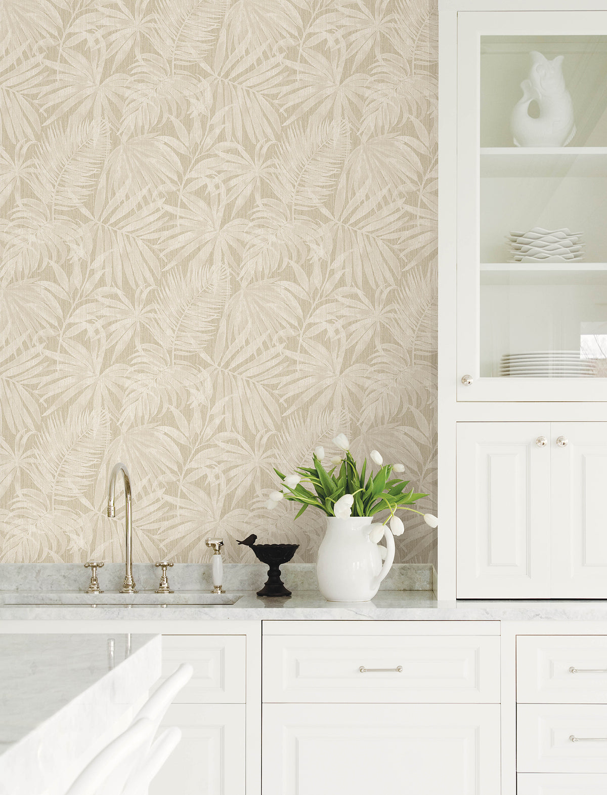 Bright kitchen with white cabinets, marble countertops, Chesapeake Sonny Fronds Wallpaper by York Wallcoverings in beige, a vase of white tulips, and a glass-front cabinet displaying plates and a decorative pitcher shaped like a bird.