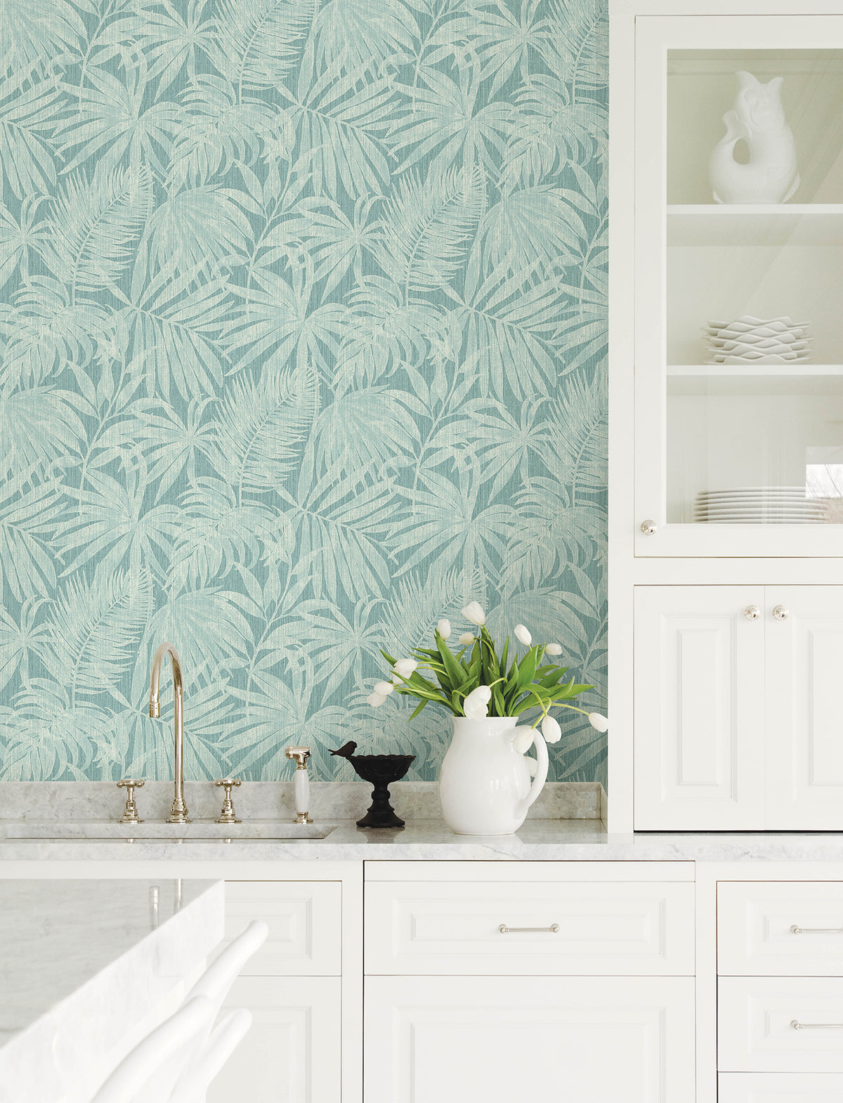 Bright white kitchen with marble countertops, crisp white cabinets, and a soothing touch from York Wallcoverings’ Chesapeake Sonny Fronds Wallpaper in light blue. Near the sink, a vase of white tulips stands beside a small bird figurine. Dishes are neatly stacked in a glass-front cabinet.