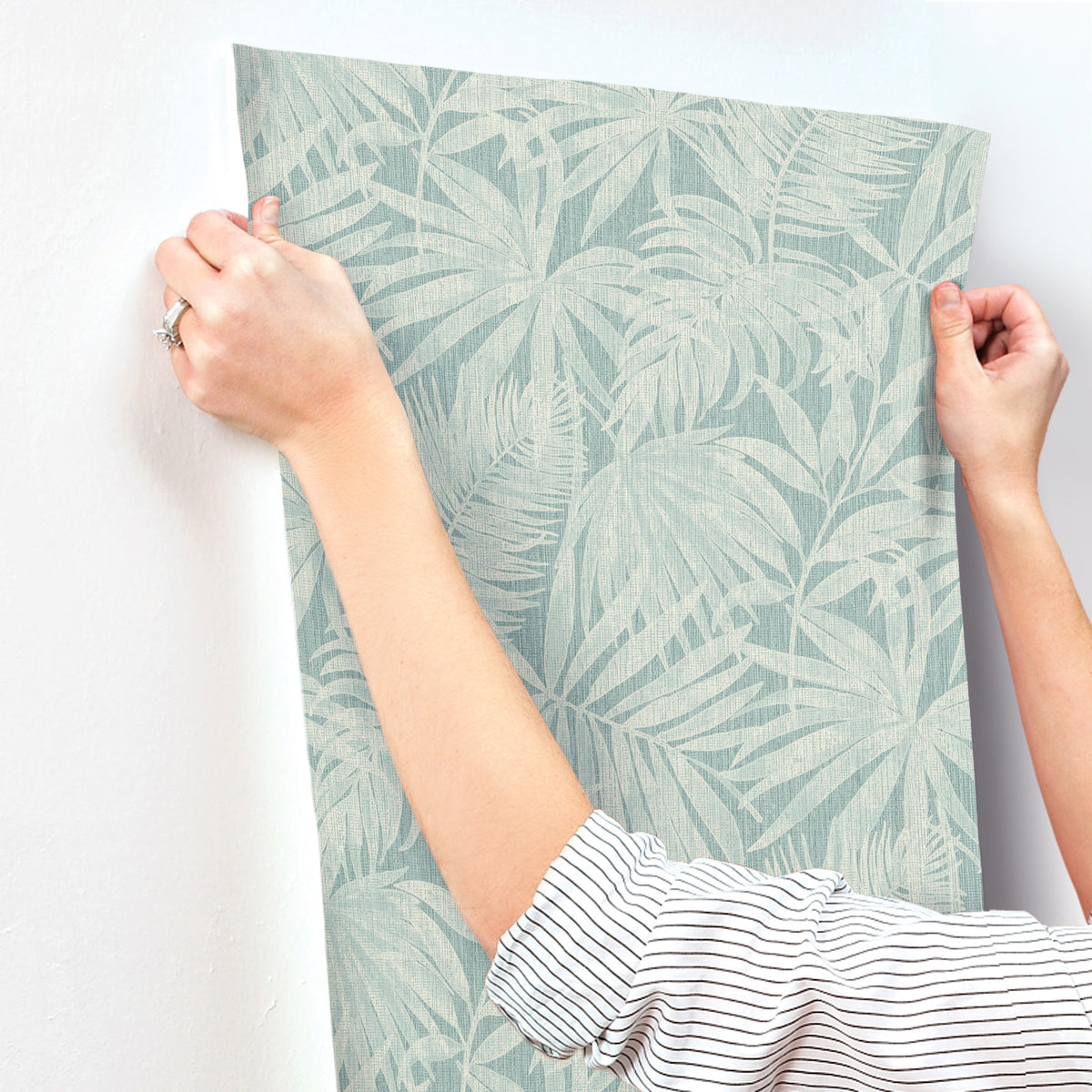 A person in a striped shirt is applying Chesapeake Sonny Fronds Wallpaper by York Wallcoverings, which features a pale blue leafy pattern, to a white wall.