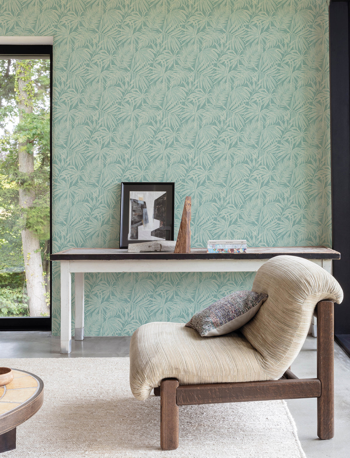 A modern living space featuring a textured beige chair, a table displaying framed art and decor, and walls adorned with York Wallcoverings’ Chesapeake Sonny Fronds Wallpaper in a light green palm pattern. A large window reveals green trees outside.