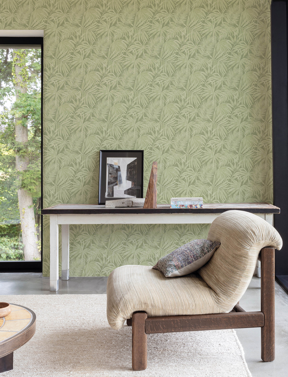 A cozy chair with a pillow sits in front of a table displaying framed art and decor, set against a green leafy-patterned wall covered in Chesapeake Sonny Fronds Wallpaper by York Wallcoverings. A large window reveals trees outside, and a light rug covers the floor.