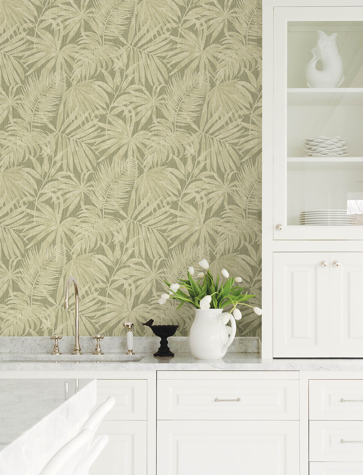 A white kitchen with marble countertops, a sink, a cabinet with glass doors, white dishes, a vase of white tulips, and York Wallcoverings’ Chesapeake Sonny Fronds Wallpaper in beige palm leaf pattern on the wall.