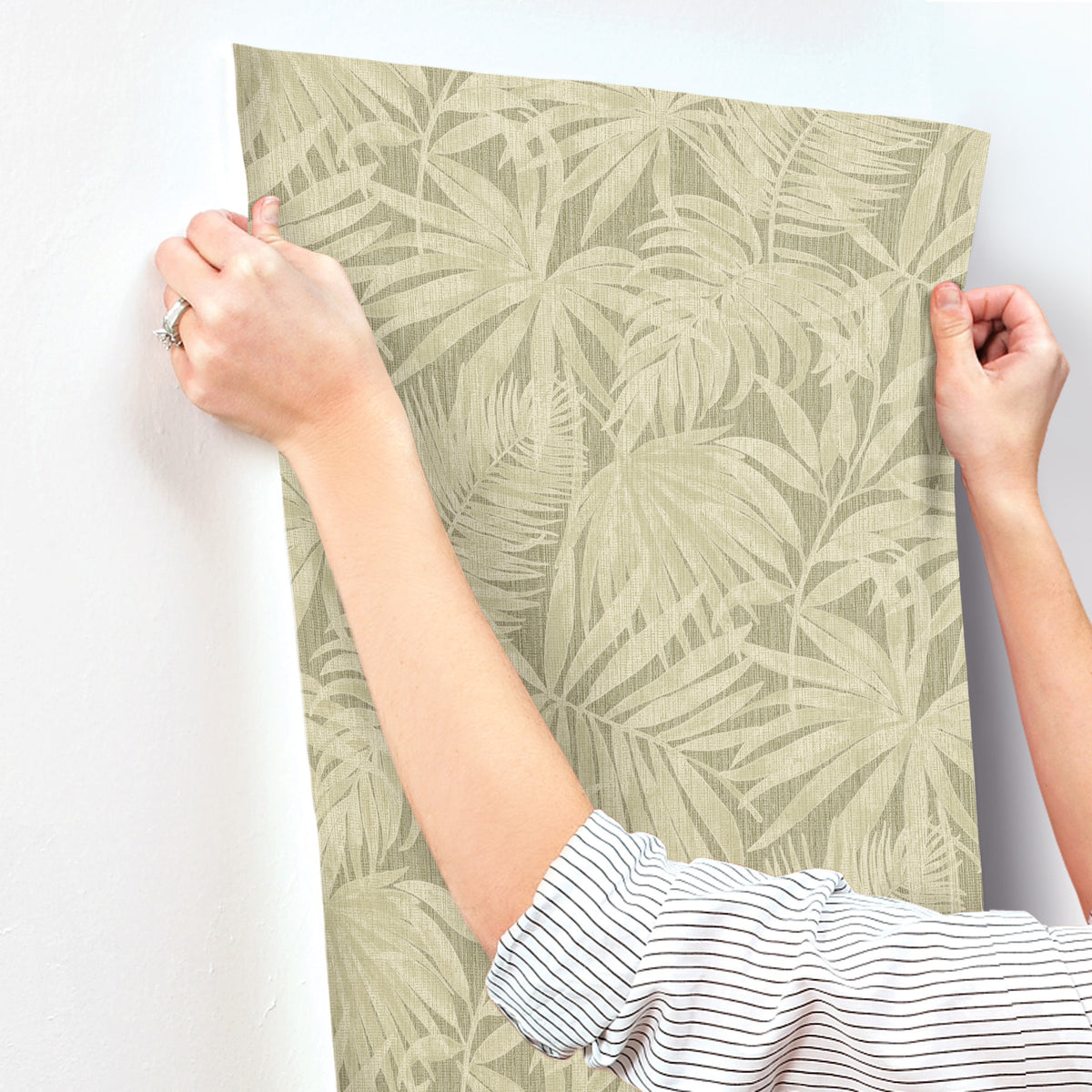 A person in a striped shirt applies Chesapeake Sonny Fronds Wallpaper by York Wallcoverings, featuring a leafy green pattern, to a white wall.