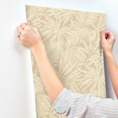 A person with a striped sleeve presses Chesapeake Sonny Fronds Wallpaper by York Wallcoverings—featuring a beige leafy pattern—onto a white wall, smoothing it with their hands.