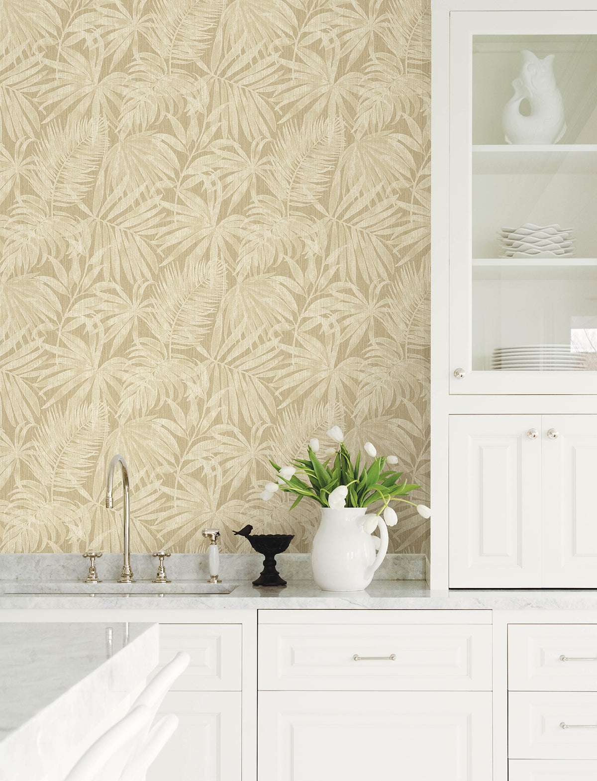 White kitchen with marble countertops, Chesapeake Sonny Fronds Wallpaper by York Wallcoverings in beige, glass-front cabinet, white ceramic pitcher with tulips, black bird decor, and chrome faucet.