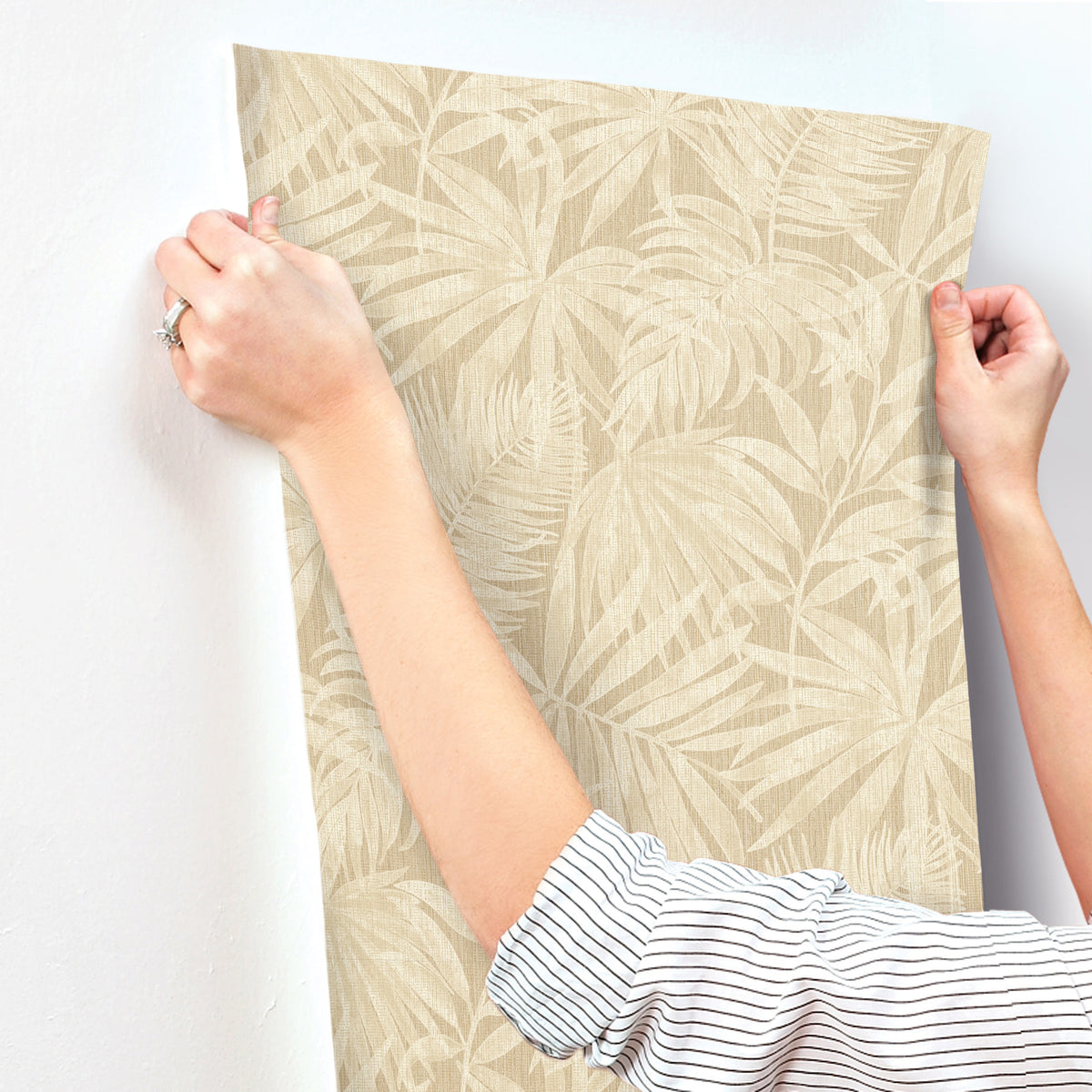 A person with a striped sleeve holds up Chesapeake Sonny Fronds Wallpaper by York Wallcoverings—featuring a beige, leaf-patterned design—against a white wall, preparing to apply it.