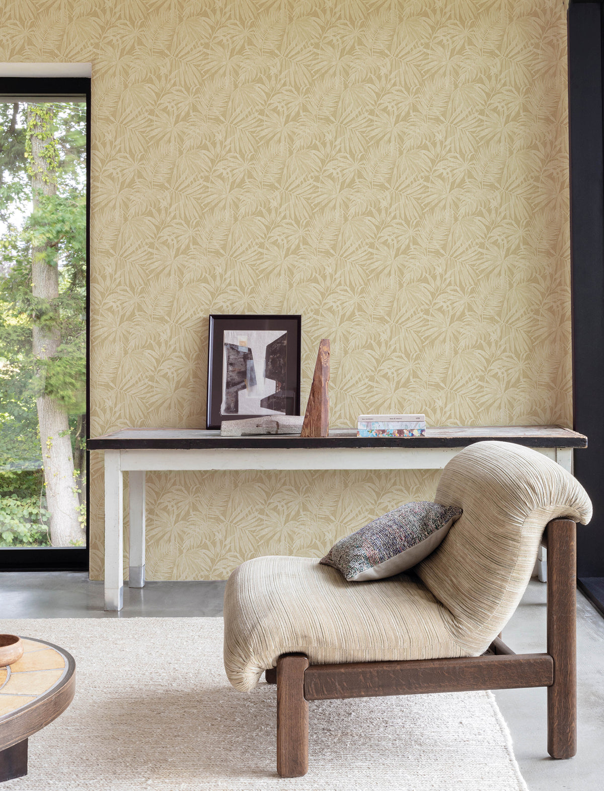 A cozy chair with a cushion sits in front of a console table displaying art and decor. The wall is adorned with Chesapeake Sonny Fronds Wallpaper by York Wallcoverings, featuring a beige, leafy design. A large window reveals green trees outside.