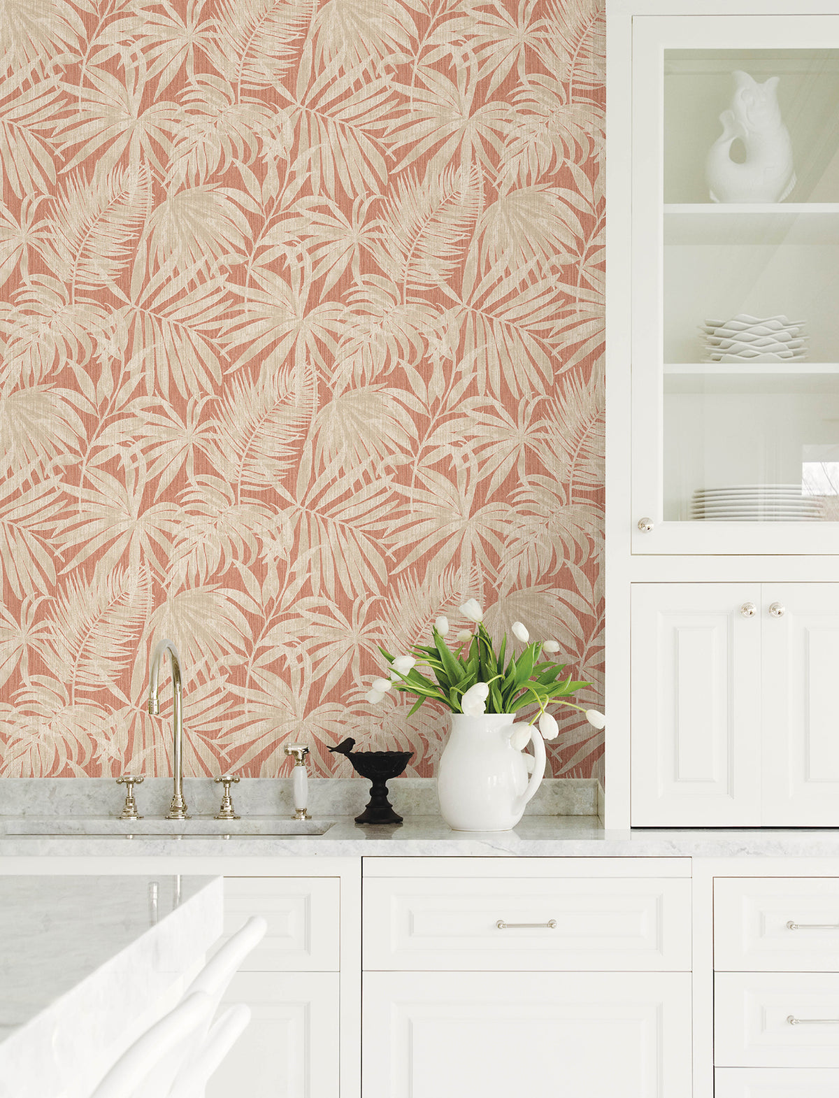 Certainly! Here’s a revised version of your sentence, incorporating the product data: Bright kitchen with white cabinets, marble countertop, a white pitcher with tulips, and a small black bird sculpture. The backsplash features Chesapeake Sonny Fronds Wallpaper by York Wallcoverings in pink with a beige leafy pattern.