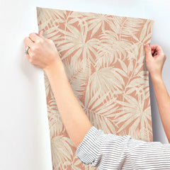 Hands are hanging a panel of York Wallcoverings Chesapeake Sonny Fronds Wallpaper, which features a beige tropical leaf pattern on a light pinkish background. The person wearing a striped long-sleeve shirt and a ring is carefully aligning the wallpaper.