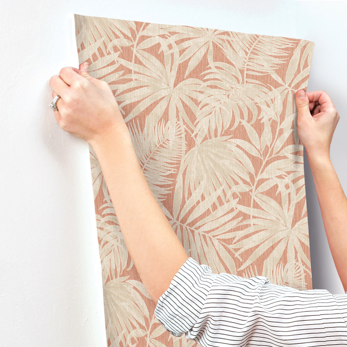 A person in a striped shirt holds up the Chesapeake Sonny Fronds Wallpaper by York Wallcoverings, featuring a beige tropical leaf pattern, against a white wall as they prepare to hang it.