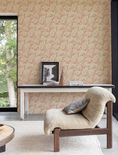 Cozy room featuring a textured beige chair, a small table with framed photos, a geometric ornament, and books set against York Wallcoverings’ Chesapeake Sonny Fronds Wallpaper. A large window reveals green trees outside.