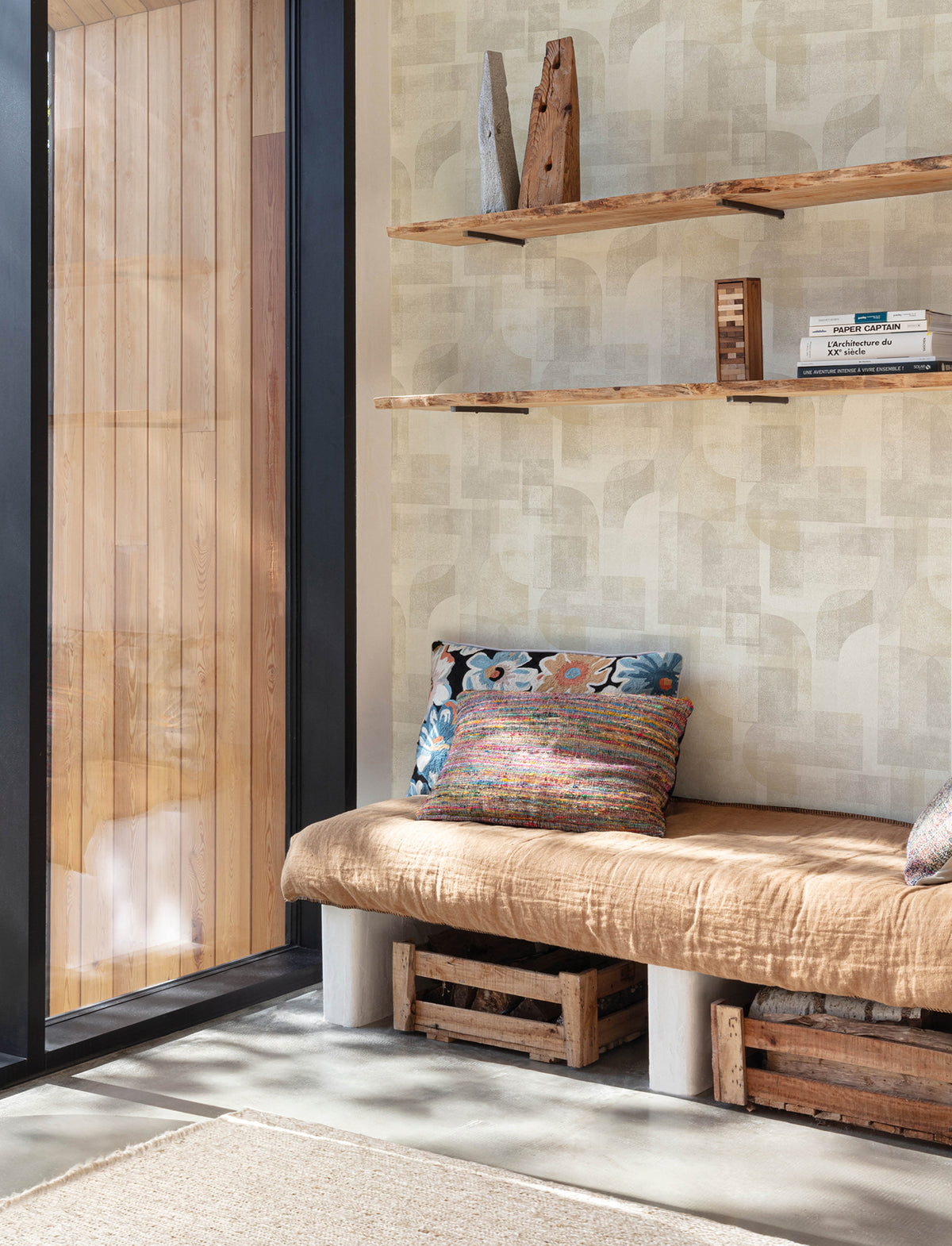 Create a cozy reading nook featuring a tan cushioned bench, colorful pillows, wooden crates for storage, and floating wooden shelves displaying books and decor—all set against the stylish Chesapeake Cassandra Block Geometric Wallpaper from York Wallcoverings. A large window fills the space with natural light.