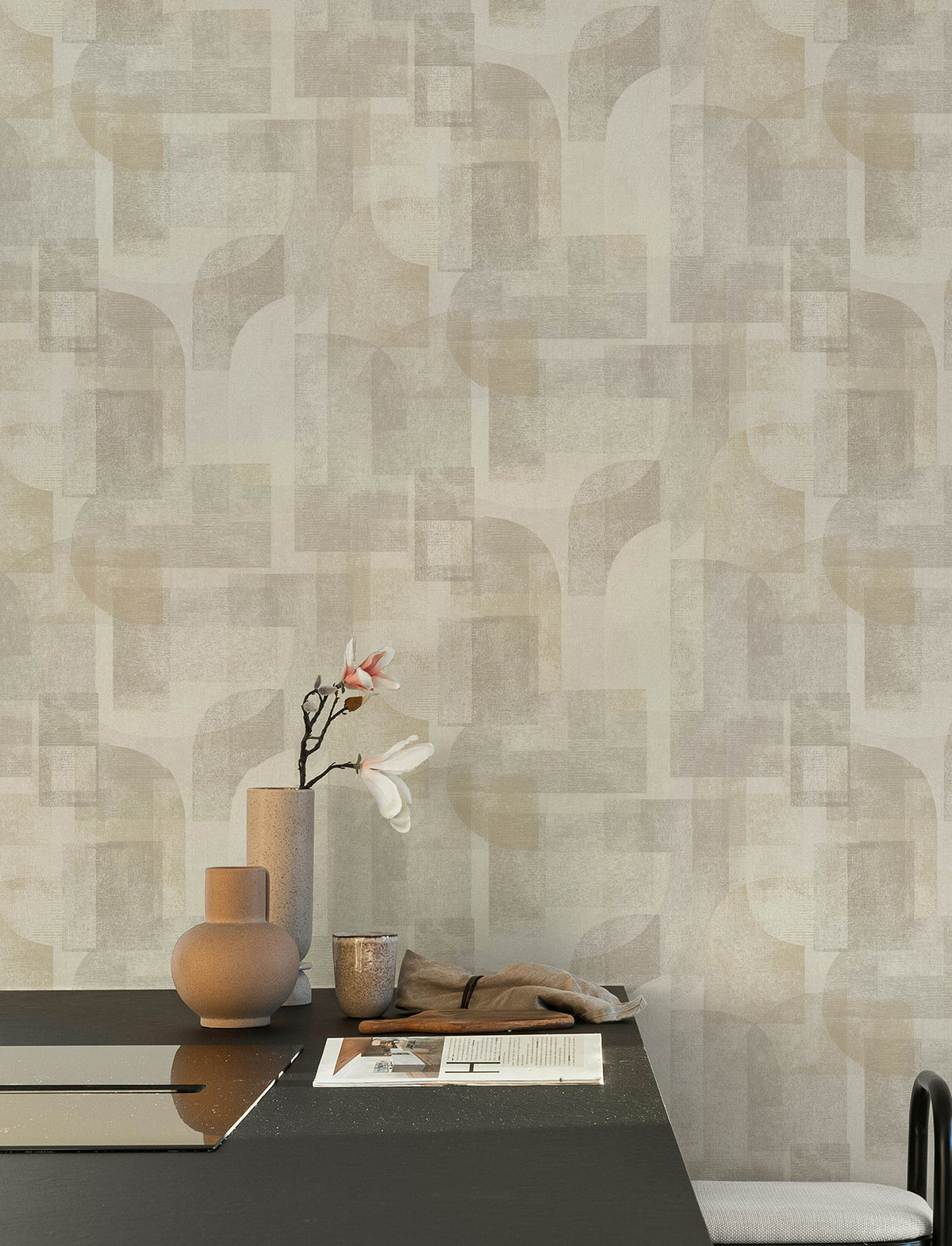 A modern table adorned with vases, a flowering branch, papers, and a cloth sits in front of a wall covered in York Wallcoverings’ Chesapeake Cassandra Block Geometric Wallpaper.