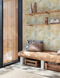 A cozy bench with colorful pillows sits next to a large window; wooden crates are used as storage underneath, and rustic shelves with books and décor hang on a wall adorned with York Wallcoverings’ Chesapeake Cassandra Block Geometric Wallpaper above.