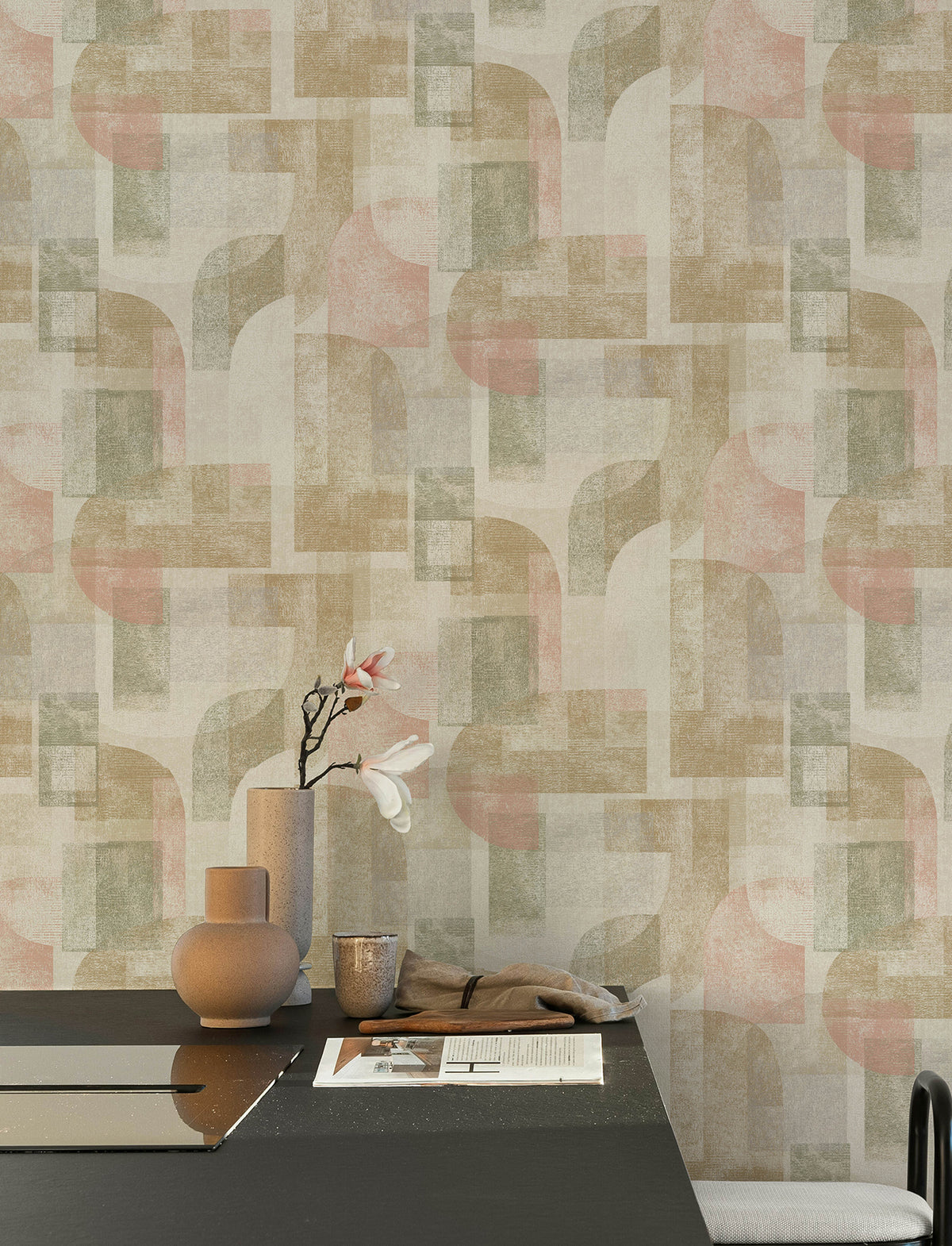 A modern dining area with a black table, beige vases, a branch with white flowers, and papers. The wall features the Chesapeake Cassandra Block Geometric Wallpaper by York Wallcoverings in muted pink, green, and beige tones.