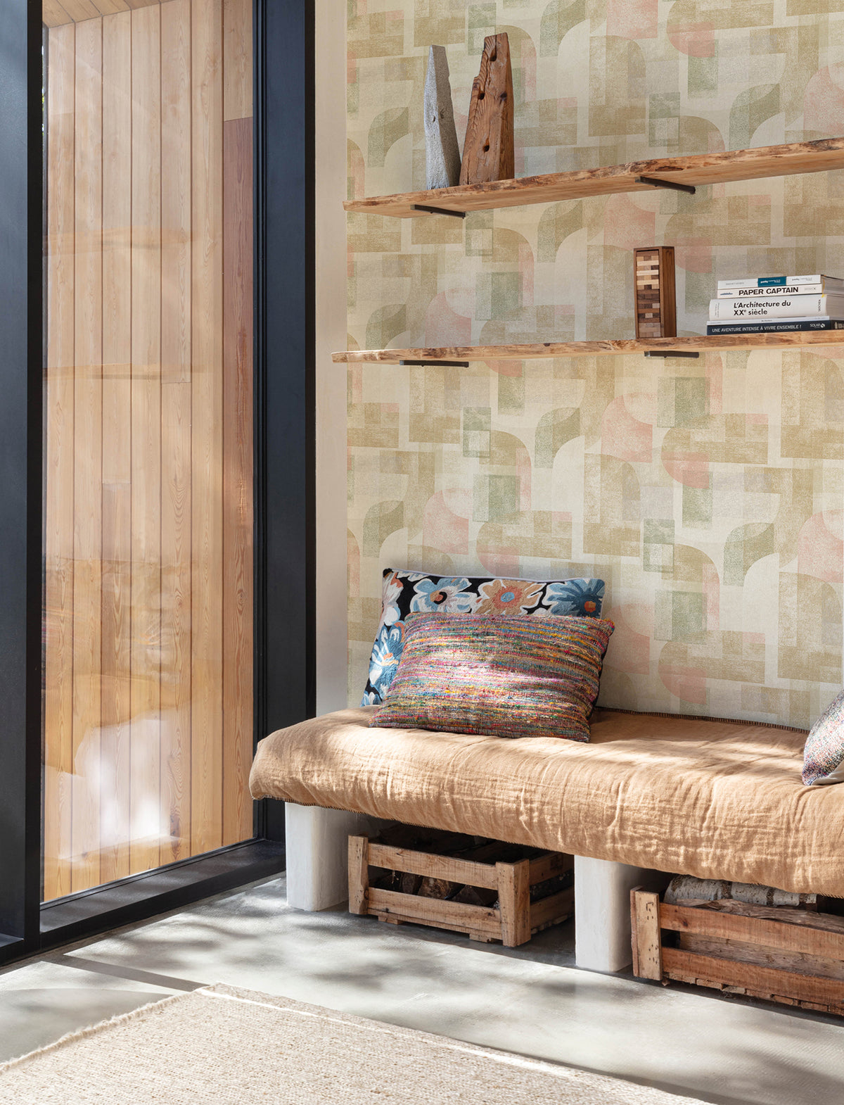 A cozy nook featuring a tan cushion bench with colorful pillows, wooden crate storage underneath, and floating shelves above displaying books and decor sits beside a large window that floods the space with natural light. The walls are adorned with York Wallcoverings’ Chesapeake Cassandra Block Geometric Wallpaper, adding a stylish retro touch.