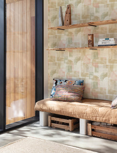 A cozy nook featuring a tan cushion bench with colorful pillows, wooden crate storage underneath, and floating shelves above displaying books and decor sits beside a large window that floods the space with natural light. The walls are adorned with York Wallcoverings’ Chesapeake Cassandra Block Geometric Wallpaper, adding a stylish retro touch.